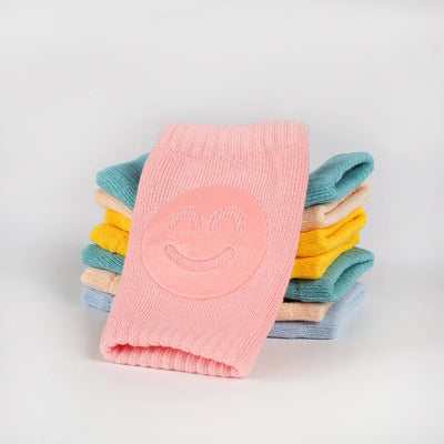 Non-slip baby knee pad in pink cotton with smiley face design, stacked with colorful pads for toddler protection during crawling.